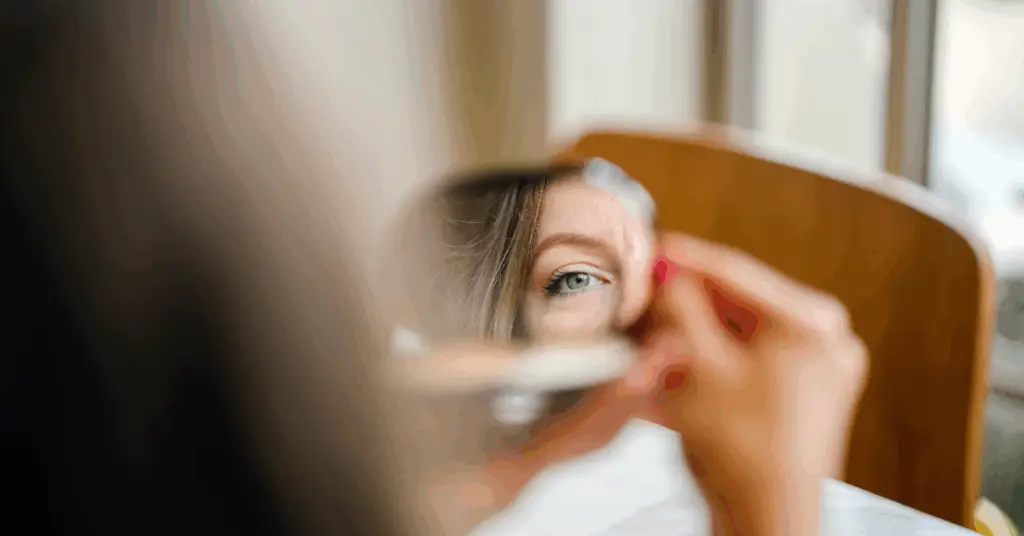 Who You Are: A woman (midlife, natural light, minimal makeup) looking into a mirror—not with vanity, but with softness or even uncertainty.